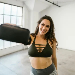 Smiling woman in gym attire holding a VR headset in a bright room.