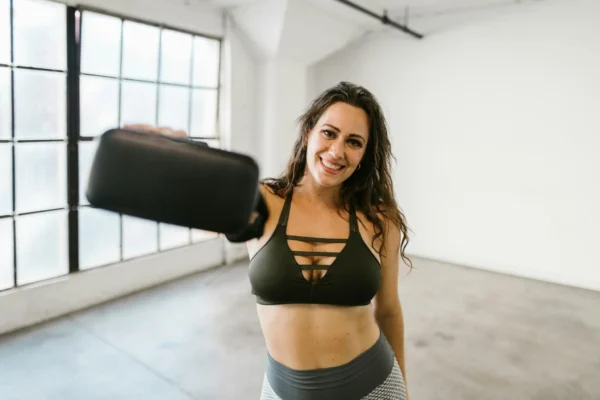 Smiling woman in gym attire holding a VR headset in a bright room.